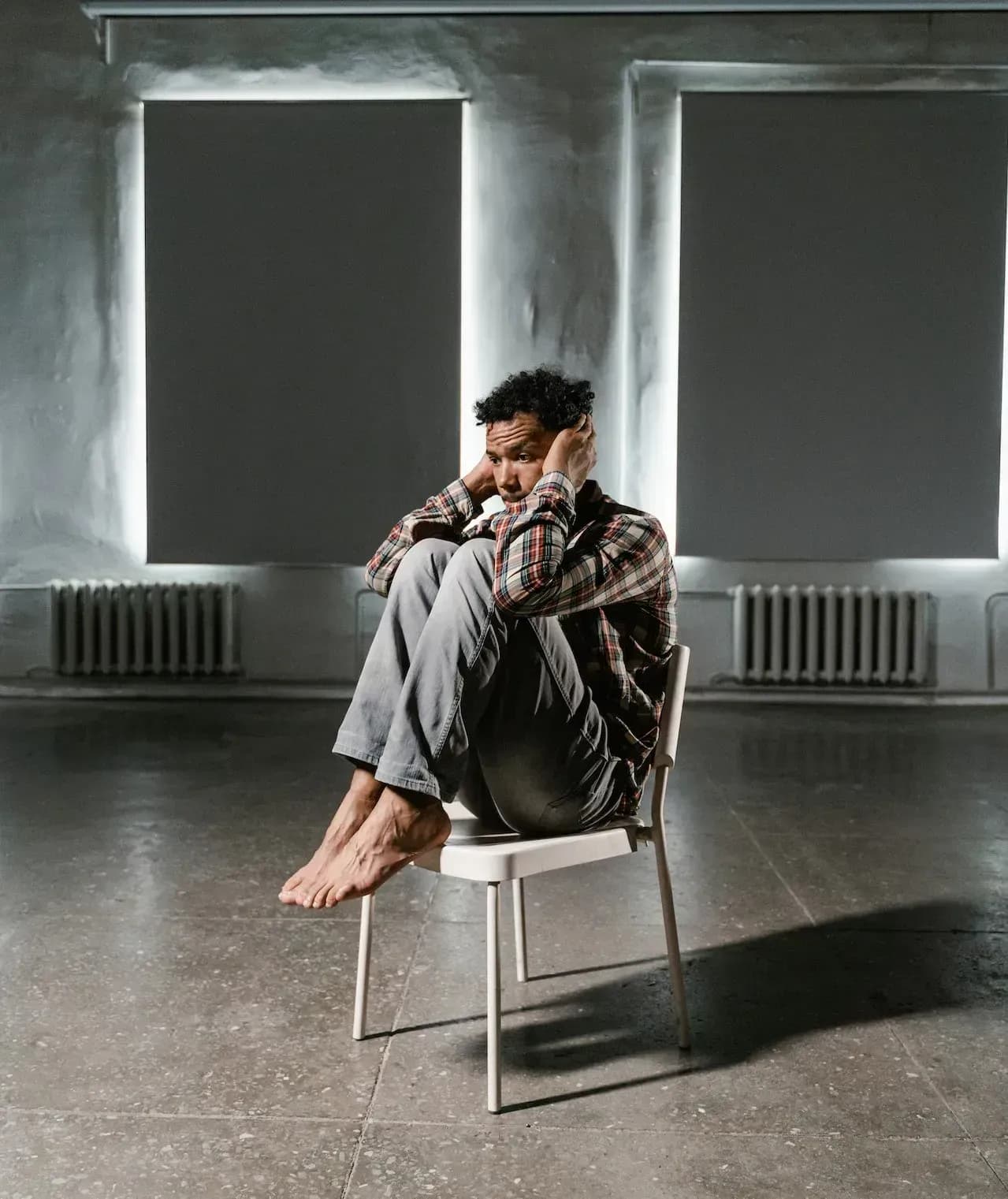 Une personne assise sur une chaise dans une pièce sombre, pensive devant de grandes fenêtres.